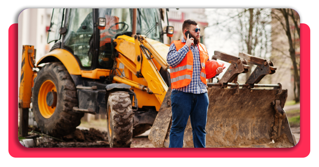 Trabajador de la construcción con casco naranja frente a un tractor con teléfono móvil en mano